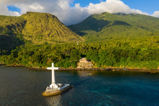 Aerial drone view of a cross in the ocean with steep Mount Vulcan and Mount Hibok-Hibok behind (Sunken Cemetery, Camiguin, Philippines)