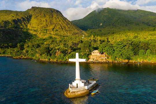 Aerial drone view of a cross in the ocean with steep Mount Vulcan and Mount Hibok-Hibok behind (Sunken Cemetery, Camiguin, Philippines)