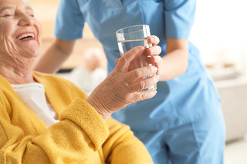 Nurse giving glass of water to senior woman indoors, closeup. Assisting elderly people