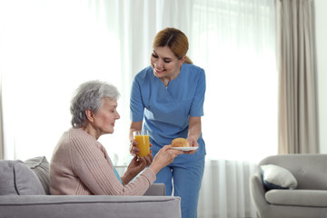 Nurse serving breakfast to elderly woman indoors, space for text. Assisting senior people