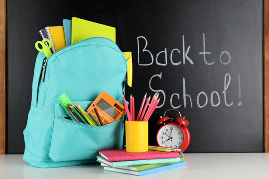 Bright Backpack With School Stationery On White Table Near Black Chalkboard. Back To School