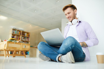 Cheerful positive hipster student guy with headphones on neck sitting with crossed legs on floor and using laptop while surfing net