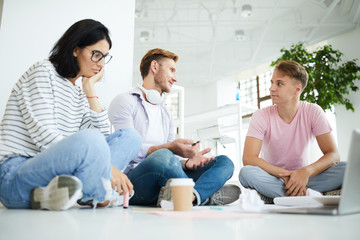 Content young people in casual clothing sitting with crossed legs on floor in spacious room and sharing ideas for project solution