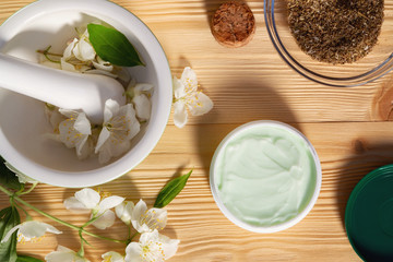Jar of cream made from natural plant ingredients, oils and herbs, jasmine flowers, mortar and pestle on a light wooden background - preparation of organic cosmetics concept. Top view, flat lay