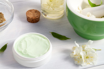 Jar of cream made from natural plant ingredients, oils and herbs, jasmine flowers, mortar and pestle on a white wooden background - preparation of organic cosmetics concept