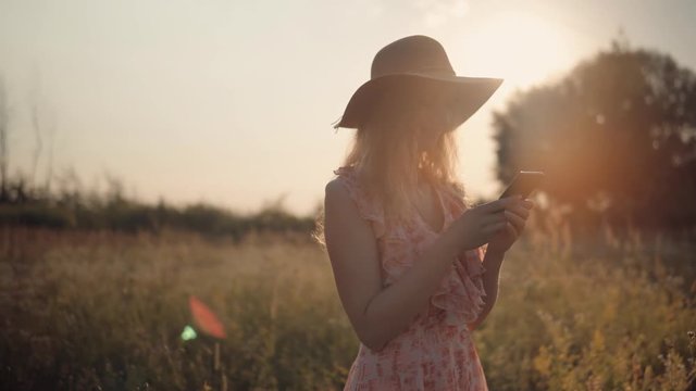 Woman In Hat Typing On Mobile At Sunset.Close Up Girl With Smartphone.Relaxed Girl Looking At Mobile Phone On Field.Beautiful Woman Having Chat Using Smartphone Outdoors.Woman Chatting With Friends.