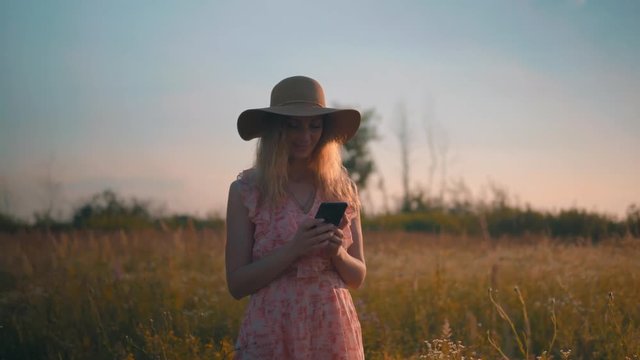 Attractive Woman Using Mobile At Sunset.Beautiful Woman Using Smartphone Outdoors.Girl In Hat Smiling And Typing On Smartphone.Woman Typing On Mobile Phone. Portrait Attractive Girl With Smatphone.