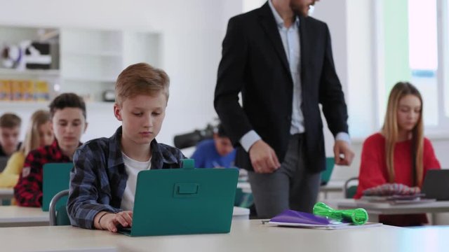 Tech young students during computer science class. Portrait of a hardworking boy pupil working on laptop with his classmates. Handsome teacher helping his kids walking in the room.