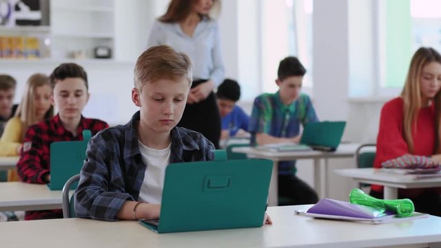 Portrait of a pretty boy pupil taking an online testing during a class. View of smart students writing a test on laptops in a computer class in modern school. Female teacher walking around and