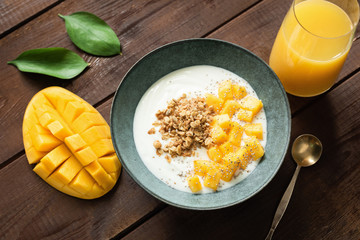 Natural yogurt with mango slices and crunchy granola in bowl and glass of orange juice on a wooden...