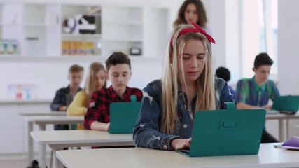 Modern education in a school. Concentrated clever teen students studying online by laptops sitting in the classroom. Pretty female teacher walking in the room monitoring a study process during class. - Powered by Adobe