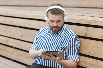 Obraz premium Serious stylish young male student listens to lectures using tablet headphones and high-speed wireless internet while sitting on a wooden park bench on a summer day. Online learning concept.