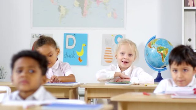 Tracking shot of cute little girl with blond hair sitting at her desk in primary school classroom and raising her hand, then answering question
