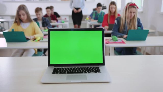 Computer science lesson in a modern school. New modern opened computer laptop with green screen on a teacher board. Female educator walking between students watching them studying online.