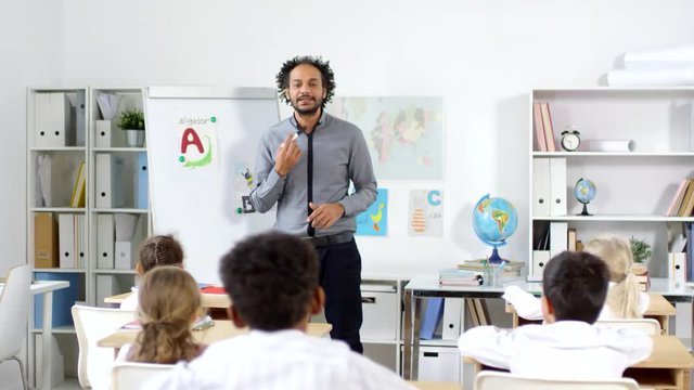 Tracking shot of male Arab preschool teacher teaching alphabet to group of little kids sitting at their desks