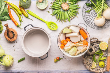 Cooking pot with spoon and white colander with fresh vegetables for vegetarian stock , broth or stew  in light kitchen table background, top view.  Copy space. Healthy food eating
