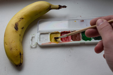 a banana watercolor paint and brush in hand of artist on white background