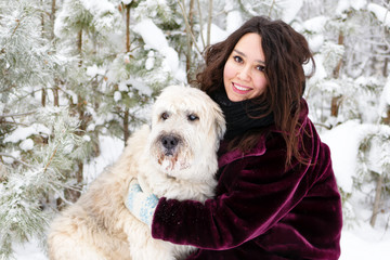 Young smiling woman is hugging her South Russian Shepherd Dog on a background of winter coniferous forest.