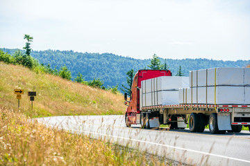 Red big rig semi truck transporting packed and tightened with slings lumber on flat bed semi...