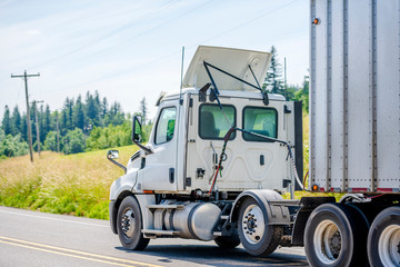 White big rig day cab semi truck with roof spoiler transporting cargo in bulk semi trailer on the road with hill with grass