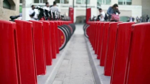 Red bicycle docks in the centre of Barcelona. Wheels and metallic parts of a bike stations. People walking around the square near museum of contemporary art. 