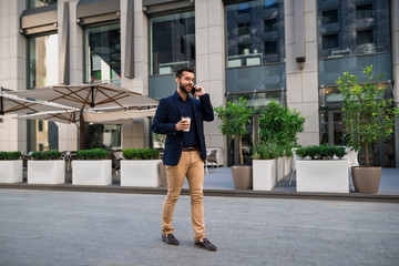 Confident young manager walking outdoors in front of cafe talking by phone and holding coffee