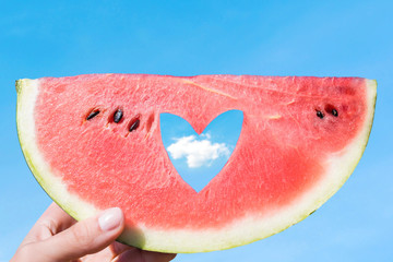 Ripe piece of watermelon with heart shape hole in female hands on the background of the blue sky with little cloud on a hot summer day. Concept