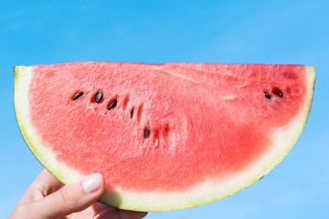 Ripe piece of watermelon in female hands on the background of the blue sky on a hot summer day. Concept
