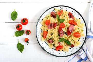 Tasty pasta farfalle with grilled sausages, fresh cherry tomatoes and basil on a plate on a white wooden background. Top view, flat lay.