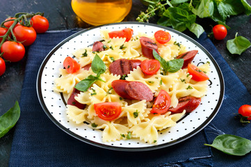 Tasty pasta farfalle with grilled sausages, fresh cherry tomatoes and basil on a plate on a black background.