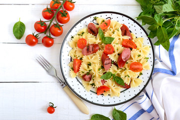 Tasty pasta farfalle with grilled sausages, fresh cherry tomatoes and basil on a plate on a white wooden background. Top view, flat lay.