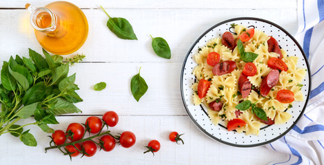 Tasty pasta farfalle with grilled sausages, fresh cherry tomatoes and basil on a plate on a white wooden background. Top view, flat lay. Banner
