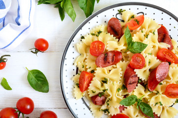 Tasty pasta farfalle with grilled sausages, fresh cherry tomatoes and basil on a plate on a white wooden background. Top view, flat lay.