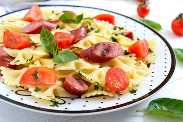 Tasty pasta farfalle with grilled sausages, fresh cherry tomatoes and basil on a plate on a white wooden background.