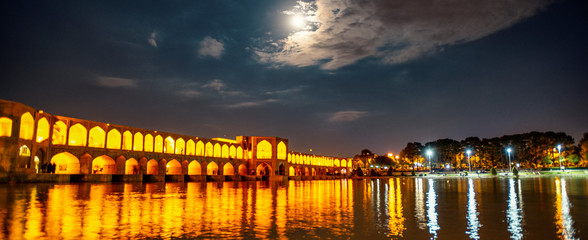 Die Khaju-Brücke über dem Zayandeh-Fluss wird in der Abenddämmerung mit Lichtern und Mond am Himmel beleuchtet und dient auch als Damm