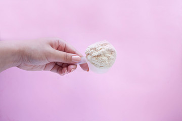 female hand holding a measuring spoon of whey protein powder with plastic spoon on pink background top view