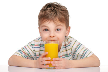 Portrait of happy little boy with glass of orange juice sitting at table on white background. Child is drinking orange juice and looking at camera.