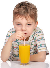 Portrait of happy little boy with glass of orange juice sitting at table on white background. Child is drinking orange juice using straw.