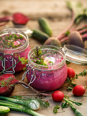 Cold beetroot soup, cold served with the addition of cucumbers, radishes, boiled eggs and fresh herbs, dill, green onions in glass jars on a wooden table. Chilled soup, excellent dish for summer