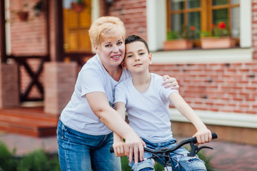 Loving mother help her cute grandson ride a bicycle near garden. Family photo.