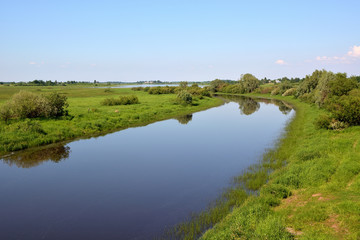 Fototapeta na wymiar Summer rural landscape with a calm river with a reflection of trees and blue sky in the water with green fields with trees and a village on the banks.River Veriaja Novgorod region