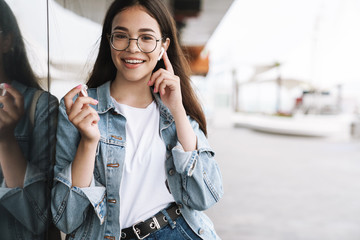 Happy young pretty woman student wearing eyeglasses walking outdoors resting listening music with...