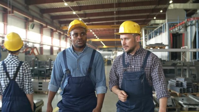 Dollying medium two shot of Caucasian and Afro-American factory employees in hardhats, walking together along factory floor with rows of metalwork components and machinery and chatting animatedly