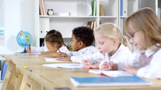 Side view shot with PAN of cute little children sitting at their desks in primary school classroom and doing writing exercises