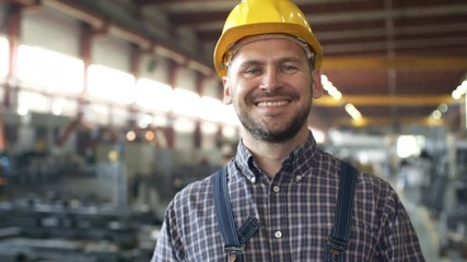 Pan slow motion close-up shot of male Caucasian factory engineer turning to camera and smiling with satisfaction, standing in sprawling sunlit workshop with blurred machinery in background - Powered by Adobe