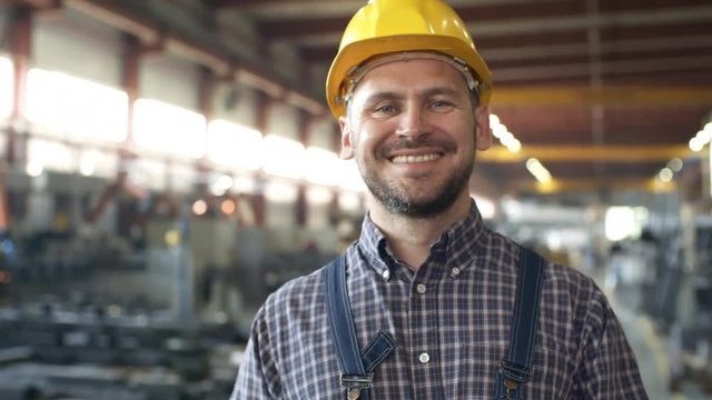 Pan slow motion close-up shot of male Caucasian factory engineer turning to camera and smiling with satisfaction, standing in sprawling sunlit workshop with blurred machinery in background