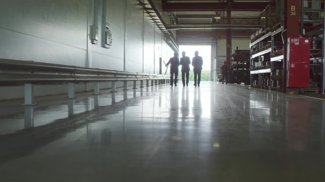 Extreme long slow motion backlit shot of three engineers wearing suits and white hardhats walking towards camera along spacious factory warehouse discussing production issues