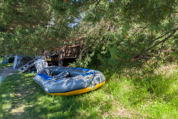 Tent camping in the shade under the trees with a wooden house and a large inflatable rafting boat near the river in the Altai Mountains. Extreme sports and recreation.
