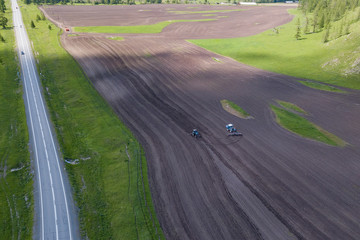 Aerial view of a farm tractor in a field during plowing of land for growing food, vegetables and fruits around the green tree with a road. The industry of production of products.