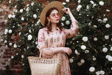 Outdoor portrait of young beautiful fashionable woman wearing wide brim straw hat, pink dress,...